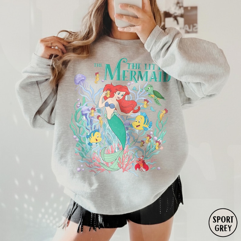 Puede incluir: Una sudadera gris claro con un gr&aacute;fico colorido de Ariel de La Sirenita. El dise&ntilde;o incluye a Ariel, peces, algas marinas y el texto "THE LITTLE MERMAID" en color turquesa. La sudadera est&aacute; etiquetada como "SPORT GREY" en un c&iacute;rculo.