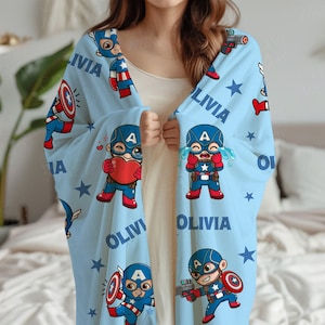 Pode incluir: Um cobertor azul claro com ilustra&ccedil;&otilde;es de desenhos animados do Capit&atilde;o Am&eacute;rica, estrelas e o nome "Olivia". O cobertor apresenta figuras do Capit&atilde;o Am&eacute;rica em v&aacute;rias poses, segurando um escudo, uma arma e um cora&ccedil;&atilde;o.