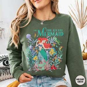 Puede incluir: Sudadera verde oliva con un gr&aacute;fico colorido de Ariel de La Sirenita. El dise&ntilde;o incluye a Ariel, Flounder, Sebasti&aacute;n y otras criaturas marinas. El texto "THE LITTLE MERMAID" est&aacute; impreso encima del gr&aacute;fico.
