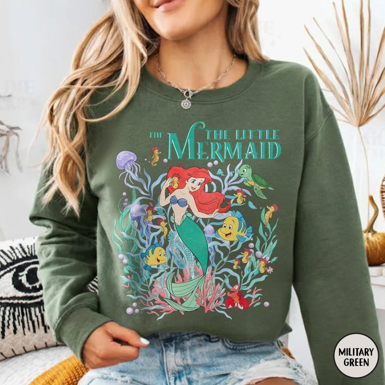 Puede incluir: Sudadera verde oliva con un gr&aacute;fico colorido de Ariel de La Sirenita. El dise&ntilde;o incluye a Ariel, Flounder, Sebasti&aacute;n y otras criaturas marinas. El texto "THE LITTLE MERMAID" est&aacute; impreso encima del gr&aacute;fico.
