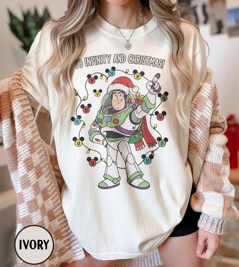 May include: Ivory-coloured t-shirt with a graphic of Buzz Lightyear in a Santa hat, encircled by Mickey Mouse-shaped Christmas lights. The text "TO INFINITY AND CHRISTMAS!" is printed above the graphic. This festive shirt is part of a holiday collection.
