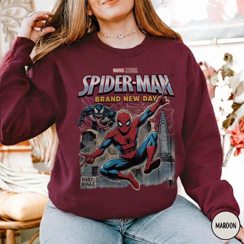 K&ouml;nnte beinhalten: Burgunderfarbenes Sweatshirt mit einem Marvel Studios Spider-Man-Motiv. Das Design zeigt Spider-Man, Venom und den Text "BRAND NEW DAY" vor einer Stadtkulisse. Das Daily Bugle-Logo ist ebenfalls zu sehen.