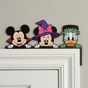 Op de afbeelding: Een decoratieve deurtopper met drie stripfiguren: Mickey Mouse verkleed als vampier met een rode cape, Minnie Mouse met een paarse heksenhoed met een roze strik, en Donald Duck als een groen monster met bouten op zijn hoofd.