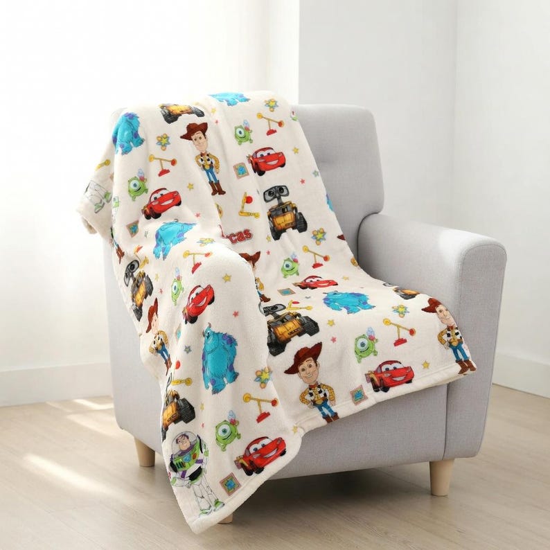 May include: A cream-colored blanket featuring colorful cartoon characters, including Woody, Buzz Lightyear, and Lightning McQueen. The blanket is draped over a gray armchair, showcasing a soft, plush texture. The blanket is decorated with various cartoon characters.