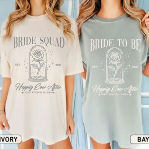 May include: Two t-shirts, one ivory and one bay, with the phrases "BRIDE SQUAD" and "BRIDE TO BE". Each shirt has a rose in a glass dome, the words "Happily Ever After" and "Magic Kingdom, Florida" printed on the front.