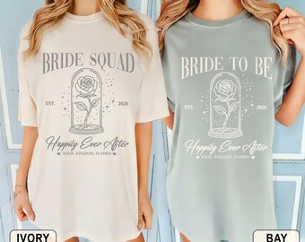 May include: Two t-shirts, one ivory and one bay, featuring the text "BRIDE SQUAD" and "BRIDE TO BE" respectively. Each shirt displays a rose in a glass dome, the words "Happily Ever After" and "Magic Kingdom, Florida."