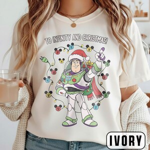 May include: Ivory t-shirt with a Buzz Lightyear graphic wearing a Santa hat, surrounded by Christmas lights with Mickey Mouse heads. The text "TO INFINITY AND CHRISTMAS!" is printed above the graphic.