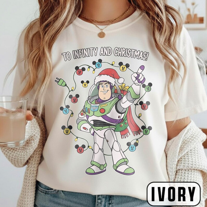 May include: Ivory t-shirt with a Buzz Lightyear graphic wearing a Santa hat, surrounded by Christmas lights with Mickey Mouse heads. The text "TO INFINITY AND CHRISTMAS!" is printed above the graphic.