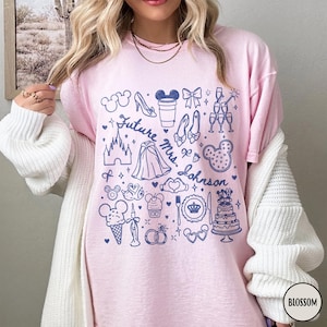 May include: A light pink t-shirt featuring a dark blue graphic print of various icons including a castle, a wedding cake, rings, and the text Future Mrs. Johnson. The design includes small stars, hearts, and bows scattered among the illustrations.
