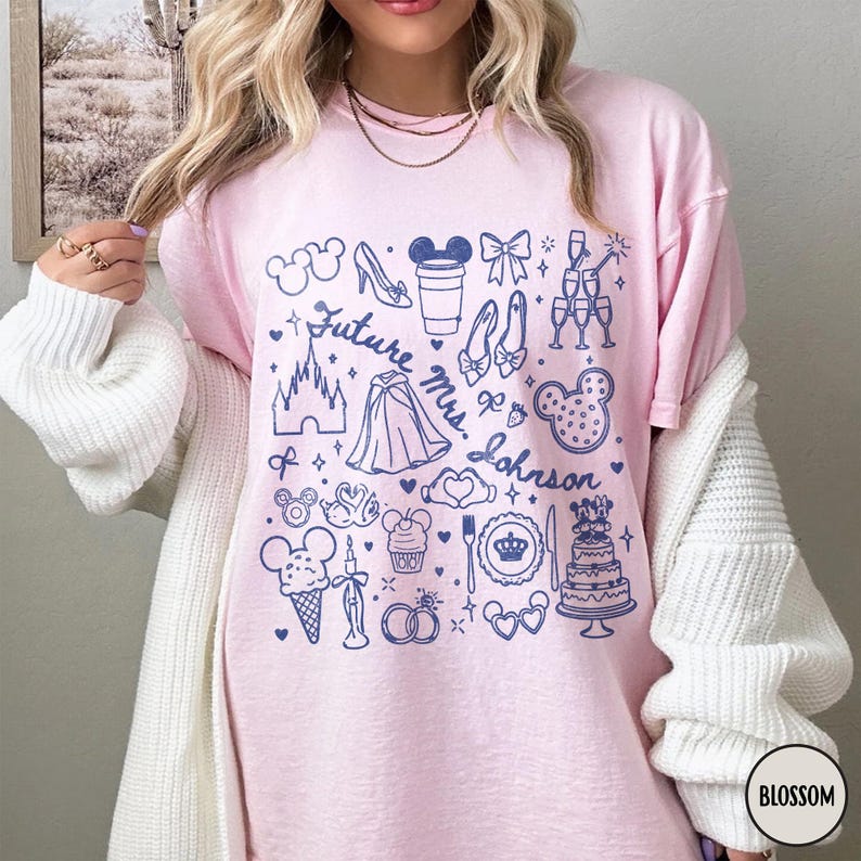 May include: A light pink t-shirt featuring a dark blue graphic print of various icons including a castle, a wedding cake, rings, and the text Future Mrs. Johnson. The design includes small stars, hearts, and bows scattered among the illustrations.