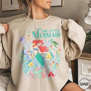 Puede incluir: Sudadera color arena con un gr&aacute;fico colorido de Ariel de La Sirenita. El dise&ntilde;o incluye a Ariel, Flounder, Sebasti&aacute;n y otras criaturas marinas, con el texto "The Little Mermaid" en verde azulado.