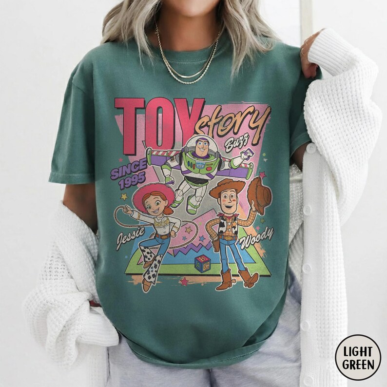 May include: Light green t-shirt with a graphic of Toy Story characters Buzz Lightyear, Woody, and Jessie. The design includes the text "Toy Story" and "Since 1995". The shirt has a crew neck and short sleeves.