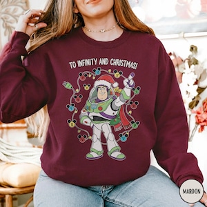 May include: Maroon sweatshirt with a graphic of Buzz Lightyear wearing a Santa hat, encircled by Mickey Mouse Christmas lights. The text "TO INFINITY AND CHRISTMAS!" is printed above the image. The sweatshirt has a crew neck and long sleeves.