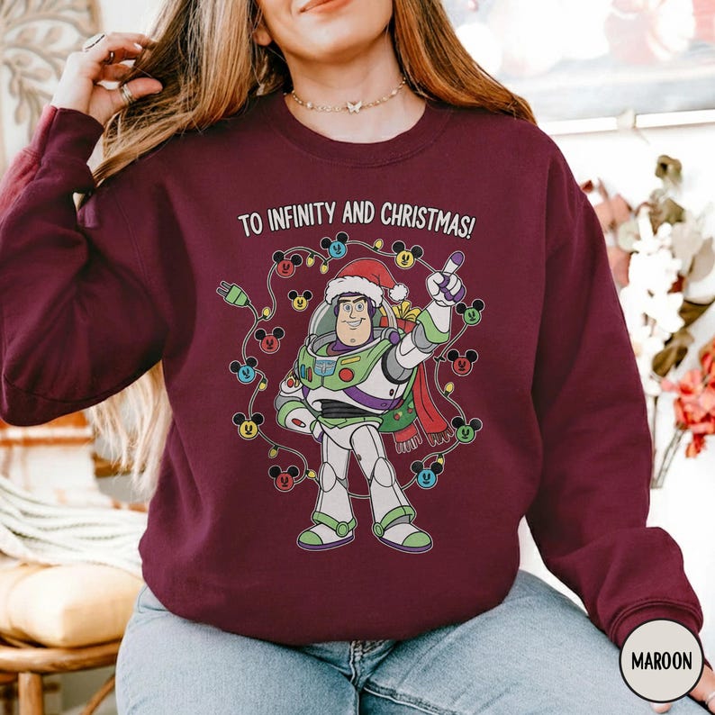 May include: Maroon sweatshirt with a graphic of Buzz Lightyear wearing a Santa hat, encircled by Mickey Mouse Christmas lights. The text "TO INFINITY AND CHRISTMAS!" is printed above the image. The sweatshirt has a crew neck and long sleeves.