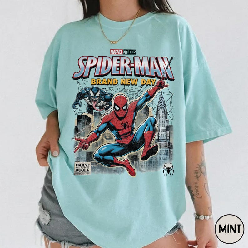 K&ouml;nnte beinhalten: Hellblaues T-Shirt mit einem Comic-Motiv von Spider-Man in Aktion, Venom, dem Daily Bugle-Logo und dem Text "Spider-Man Brand New Day". Das Marvel Studios-Logo ist ebenfalls sichtbar.