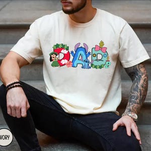 K&ouml;nnte beinhalten: Elfenbeinfarbenes T-Shirt mit dem Wort "DAD" in bunten Cartoon-Buchstaben. Das "D" ist rot mit einem hawaiianischen M&auml;dchen, das "A" ist blau mit Stitch, und das zweite "D" ist t&uuml;rkis mit einer rosa Schleife. Ein l&auml;ssiges Kleidungsst&uuml;ck.