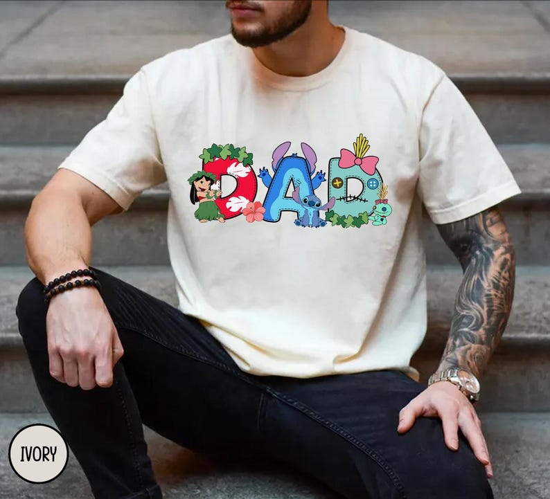 K&ouml;nnte beinhalten: Elfenbeinfarbenes T-Shirt mit dem Wort "DAD" in bunten Cartoon-Buchstaben. Das "D" ist rot mit einem hawaiianischen M&auml;dchen, das "A" ist blau mit Stitch, und das zweite "D" ist t&uuml;rkis mit einer rosa Schleife. Ein l&auml;ssiges Kleidungsst&uuml;ck.