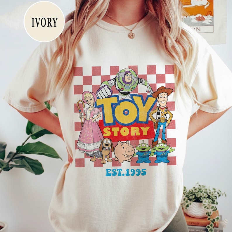 以下が含まれることがあります： アイボリー色のTシャツ。赤と白のチェック柄の背景に、ウッディやバズ・ライトイヤーなどのキャラクターが描かれた「トイ・ストーリー」のグラフィック。 「TOY STORY」の文字が大きく表示され、その下に「EST.1995」と書かれています。