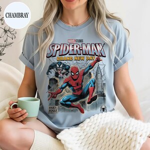 K&ouml;nnte beinhalten: Hellblaues T-Shirt mit einem Spider-Man-Motiv von Marvel Studios. Das Design zeigt Spider-Man in einer dynamischen Pose, den Text "SPIDER-MAN BRAND NEW DAY" und das Daily Bugle-Logo. Die &Auml;rmel des Shirts sind hochgekrempelt.