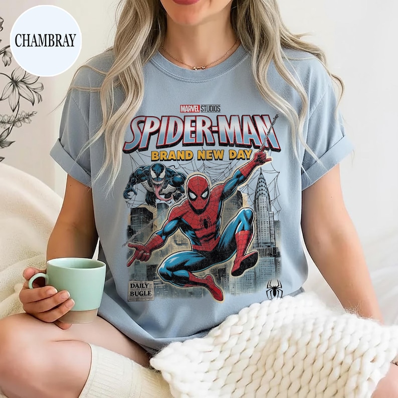 K&ouml;nnte beinhalten: Hellblaues T-Shirt mit einem Spider-Man-Motiv von Marvel Studios. Das Design zeigt Spider-Man in einer dynamischen Pose, den Text "SPIDER-MAN BRAND NEW DAY" und das Daily Bugle-Logo. Die &Auml;rmel des Shirts sind hochgekrempelt.