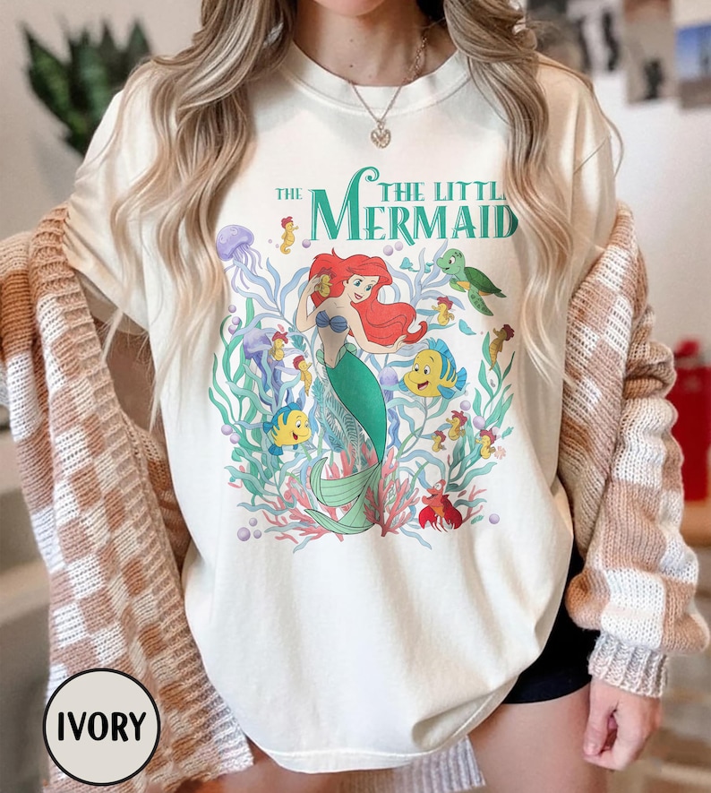 Puede incluir: Camiseta de color marfil con un gr&aacute;fico colorido de Ariel de La Sirenita. El dise&ntilde;o incluye a Ariel con cabello rojo y cola verde, rodeada de criaturas marinas y el texto "The Little Mermaid".