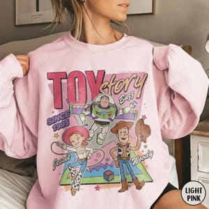 May include: Pale pink sweatshirt with a graphic print of Toy Story characters, including Buzz Lightyear, Woody, and Jessie. The design incorporates the text "TOY STORY" and "SINCE 1995". The sweatshirt has a crew neck.