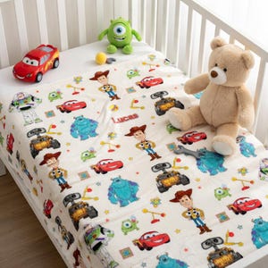 May include: A cream-colored baby blanket featuring cartoon characters from Disney Pixar movies. The blanket has a repeating pattern of characters like Woody, Buzz Lightyear, Sully, and Lightning McQueen. The name "Lucas" is printed on the blanket. A red toy car, a green monster plush, and a teddy bear are also present.