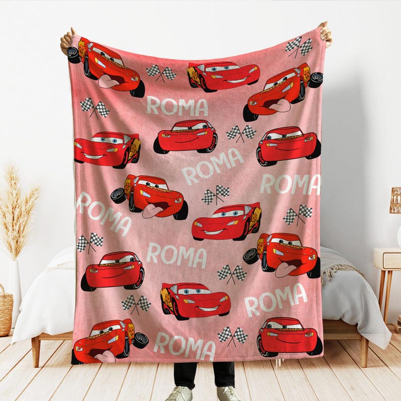 Puede incluir: Una manta rosa y roja con un patr&oacute;n repetido de un coche de carreras de dibujos animados rojo, banderas a cuadros y la palabra "ROMA". La manta est&aacute; sujeta por una persona.