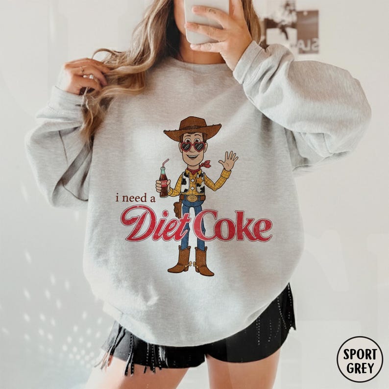 K&ouml;nnte beinhalten: Hellgraues Sweatshirt mit einem Cartoon von Woody, der eine Diet Coke-Flasche h&auml;lt. Der Text "I need a Diet Coke" ist unter der Figur in roter Schrift gedruckt. Das Sweatshirt hat lange &Auml;rmel und einen Rundhalsausschnitt.