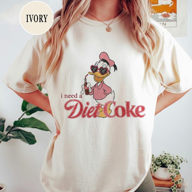 K&ouml;nnte beinhalten: Elfenbeinfarbenes T-Shirt mit einer Cartoon-Ente, die eine herzf&ouml;rmige Sonnenbrille und ein rosa Outfit tr&auml;gt und aus einer Diet Coke-Flasche trinkt. Der Text "i need a Diet Coke" ist in einer Retro-Schriftart gedruckt.