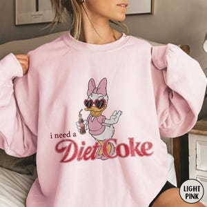 K&ouml;nnte beinhalten: Hellrosa Sweatshirt mit einem Daisy Duck-Cartoon-Design. Daisy Duck tr&auml;gt herzf&ouml;rmige Sonnenbrillen und h&auml;lt eine Diet Coke-Flasche. Der Text "i need a Diet Coke" ist in roter Schrift gedruckt. Das Sweatshirt hat einen Rundhalsausschnitt.