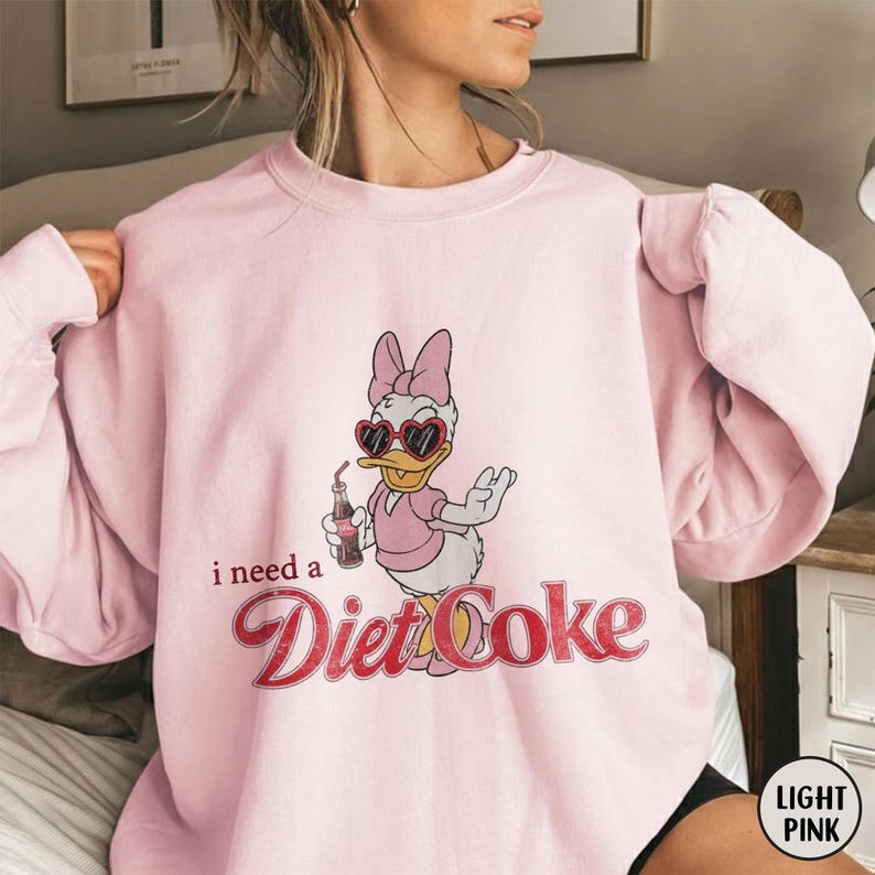 K&ouml;nnte beinhalten: Hellrosa Sweatshirt mit einem Daisy Duck-Cartoon-Design. Daisy Duck tr&auml;gt herzf&ouml;rmige Sonnenbrillen und h&auml;lt eine Diet Coke-Flasche. Der Text "i need a Diet Coke" ist in roter Schrift gedruckt. Das Sweatshirt hat einen Rundhalsausschnitt.