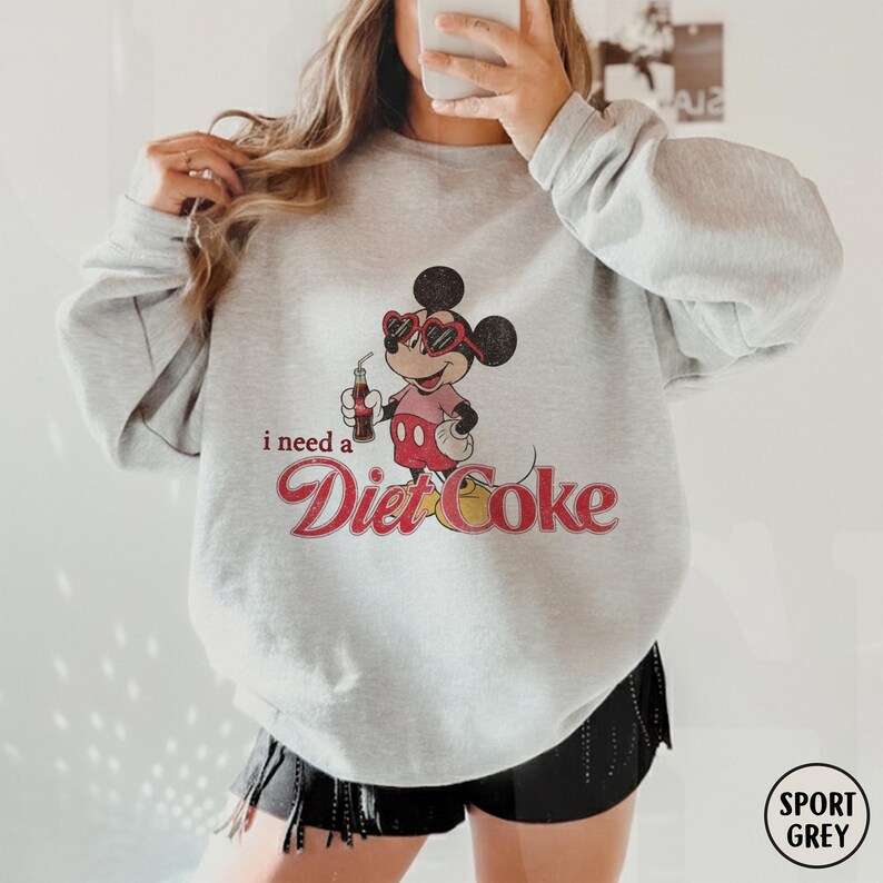 May include: Sport grey sweatshirt featuring a graphic of Mickey Mouse wearing sunglasses and holding a Diet Coke. The text "i need a Diet Coke" is printed below the graphic in a retro font. The sweatshirt is a casual style.