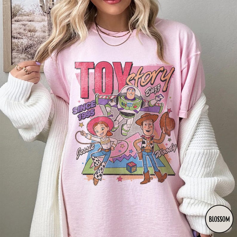 May include: Pale pink t-shirt with a graphic of Toy Story characters Buzz Lightyear, Jessie, and Woody. The shirt features the text "Toy Story" and "Since 1995". The design is in a retro style.