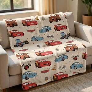Puede incluir: Una manta de color crema con dibujos de coches en rojo, azul y marr&oacute;n. La manta tiene un patr&oacute;n repetido de coches de carreras, conos de tr&aacute;fico y banderas a cuadros. La palabra "Cars" tambi&eacute;n es visible.