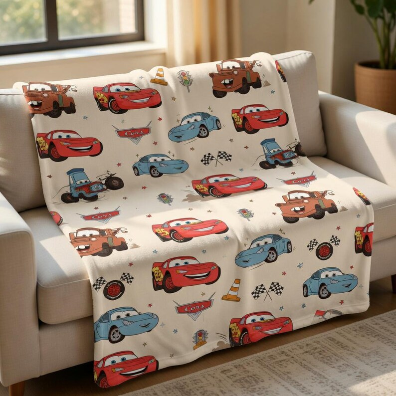 Puede incluir: Una manta de color crema con dibujos de coches en rojo, azul y marr&oacute;n. La manta tiene un patr&oacute;n repetido de coches de carreras, conos de tr&aacute;fico y banderas a cuadros. La palabra "Cars" tambi&eacute;n es visible.