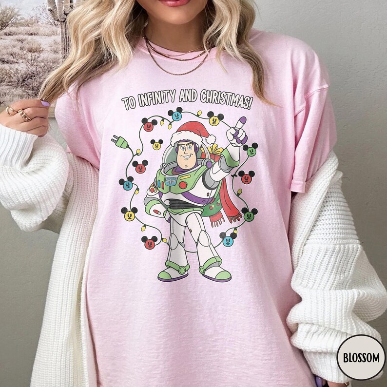 May include: Pale pink t-shirt with a Buzz Lightyear graphic wearing a Santa hat, encircled by Mickey Mouse Christmas lights. The text "TO INFINITY AND CHRISTMAS!" is printed above. The shirt has a loose fit.