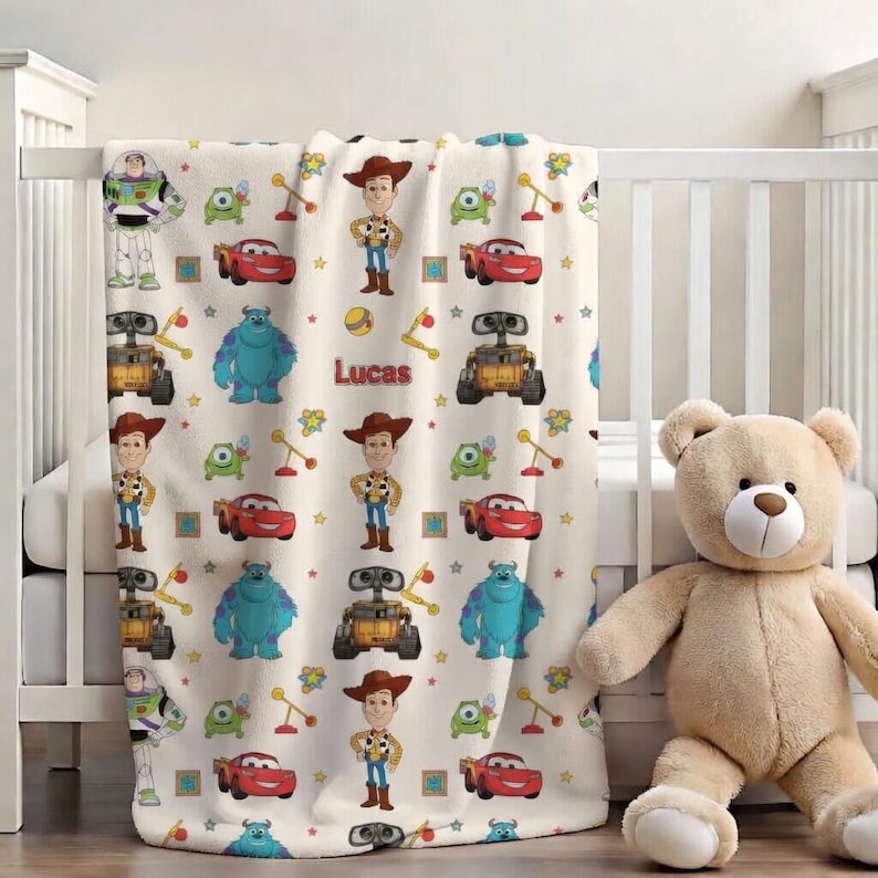 May include: A cream-colored baby blanket featuring cartoon characters from the movie Toy Story and Monsters, Inc. The blanket has images of Woody, Buzz Lightyear, Sully, and others. The name "Lucas" is printed on the blanket.
