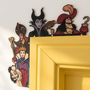 Può includere: Una decorazione angolare per porta in legno con ritagli colorati e stratificati di cinque cattivi animati. Il design include personaggi con tratti distintivi come un copricapo con corna, un cappello con piume rosse, una corona e un bastone su una cornice gialla.