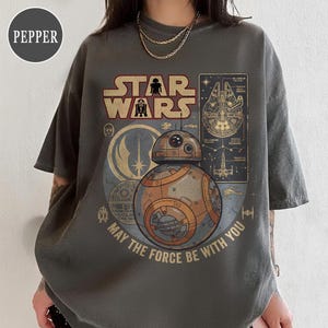 Könnte beinhalten: Dunkelgraues T-Shirt mit einem Star Wars-Motiv. Das Design zeigt das Star Wars-Logo, BB-8, den Millennium Falken und den Satz "May the Force Be With You". Die Grafik ist in Orange-, Braun- und Goldtönen gehalten.