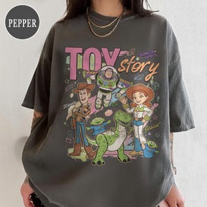 May include: Dark gray t-shirt featuring a colorful graphic of Toy Story characters, including Woody, Buzz Lightyear, and Jessie. The text "TOY STORY" is displayed in pink and orange, with "SINCE 1995" above the word "story".