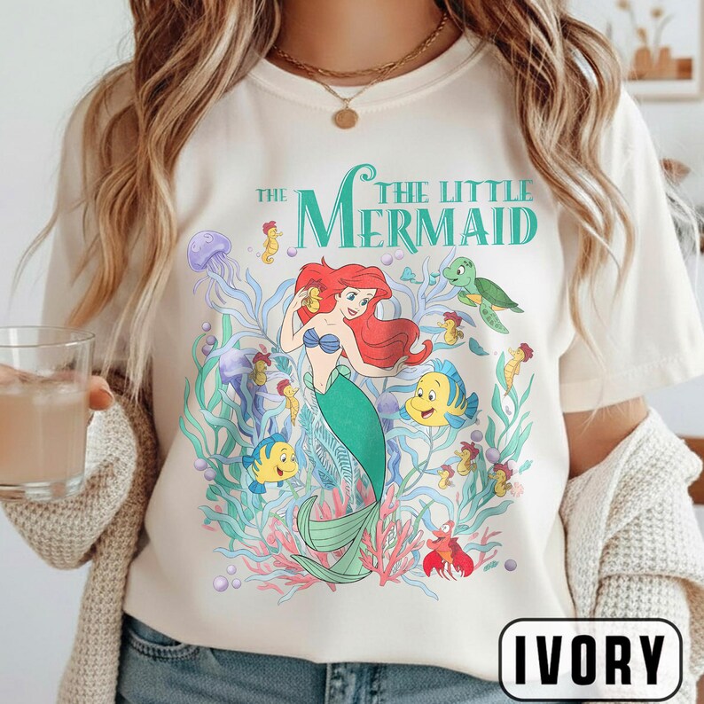 Puede incluir: Camiseta de color marfil con un gr&aacute;fico colorido de Ariel de La Sirenita. El dise&ntilde;o incluye a Ariel, Flounder, Sebasti&aacute;n y otras criaturas marinas, con las palabras "The Little Mermaid" en verde azulado.