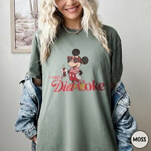 May include: Moss green t-shirt featuring a cartoon Mickey Mouse character wearing sunglasses and holding a Diet Coke bottle. The text "I need a Diet Coke" is printed in red script. The shirt is a relaxed fit.