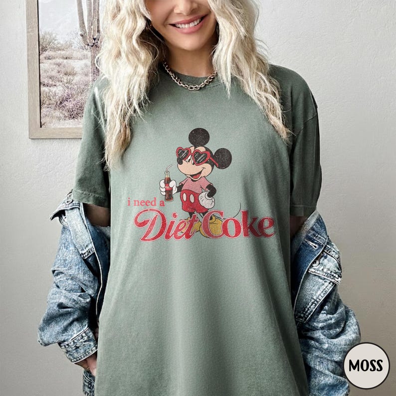 May include: Moss green t-shirt featuring a cartoon Mickey Mouse character wearing sunglasses and holding a Diet Coke bottle. The text "I need a Diet Coke" is printed in red script. The shirt is a relaxed fit.