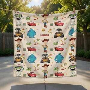 May include: A cream-colored blanket featuring cartoon characters from the movie Toy Story, including Woody, Buzz Lightyear, and others. The blanket has a repeating pattern of characters, cars, and stars, with the name "Lucas" printed in the center.
