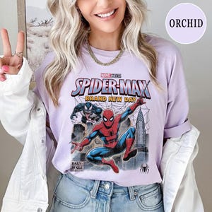 K&ouml;nnte beinhalten: Ein helllila T-Shirt mit einer Marvel Studios Spider-Man Grafik. Das Design zeigt Spider-Man in einer dynamischen Pose, den Text "SPIDER-MAN BRAND NEW DAY" und das Daily Bugle Logo. Das Shirt ist Teil der Orchid-Kollektion.
