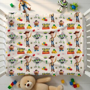 May include: A cream-colored baby blanket featuring a repeating pattern of Toy Story characters, including Woody, Buzz Lightyear, and Mr. Potato Head. The blanket also has the words "Toy Story" printed on it, along with colorful building blocks and green aliens.