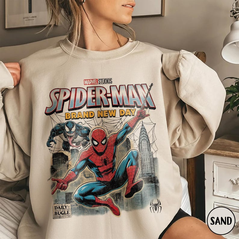 K&ouml;nnte beinhalten: Sandfarbenes Sweatshirt mit einer Grafik von Spider-Man und Venom, sowie dem Text "SPIDER-MAN BRAND NEW DAY" und "DAILY BUGLE". Das Design beinhaltet eine Stadtlandschaft und Spinnweben. Das Marvel Studios Logo ist ebenfalls sichtbar.