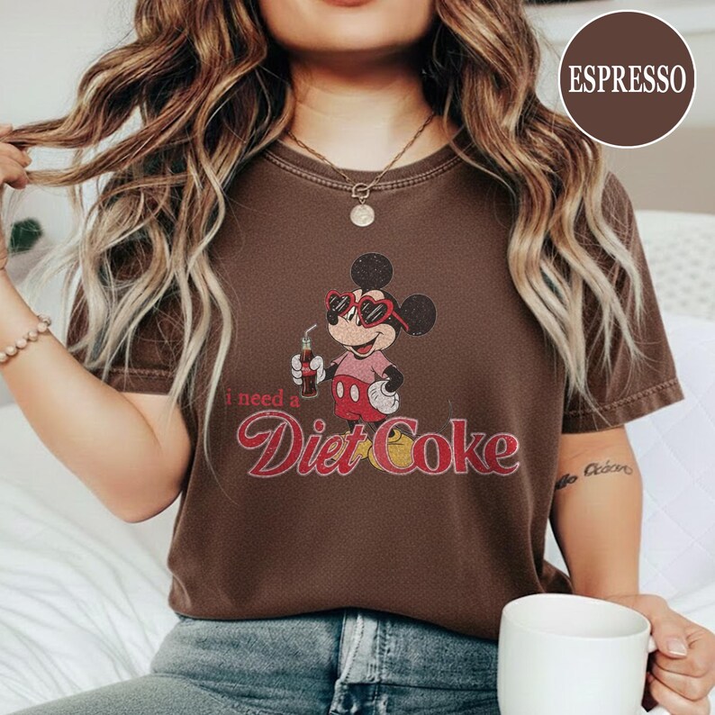 May include: Espresso-colored t-shirt featuring a graphic of Mickey Mouse wearing heart-shaped sunglasses and holding a Diet Coke. The text "I need a Diet Coke" is printed below the graphic in a retro font.