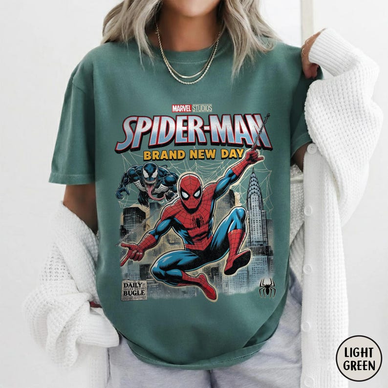 K&ouml;nnte beinhalten: Hellgr&uuml;nes T-Shirt mit einem Spider-Man-Motiv von Marvel Studios. Das Design zeigt Spider-Man, den Text "SPIDER-MAN BRAND NEW DAY" und das Daily Bugle-Logo. Eine Venom-Figur ist ebenfalls abgebildet.